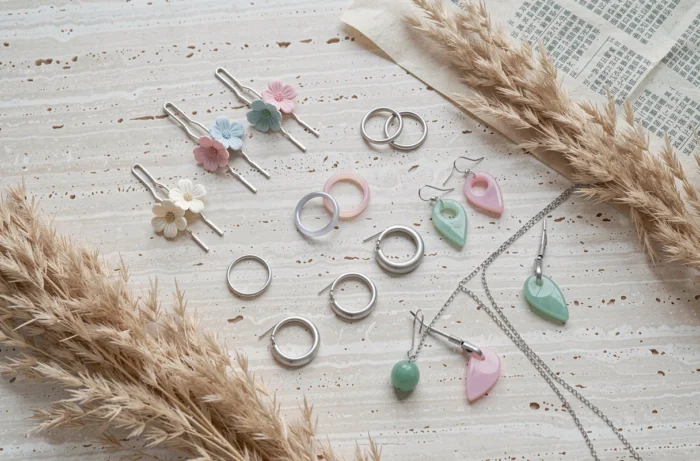 Mise à plat de bijoux d'inspiration coréenne sur marbre travertin, incluant épingles florales, bagues fines et boucles d'oreilles asymétriques en argent et résine pastel